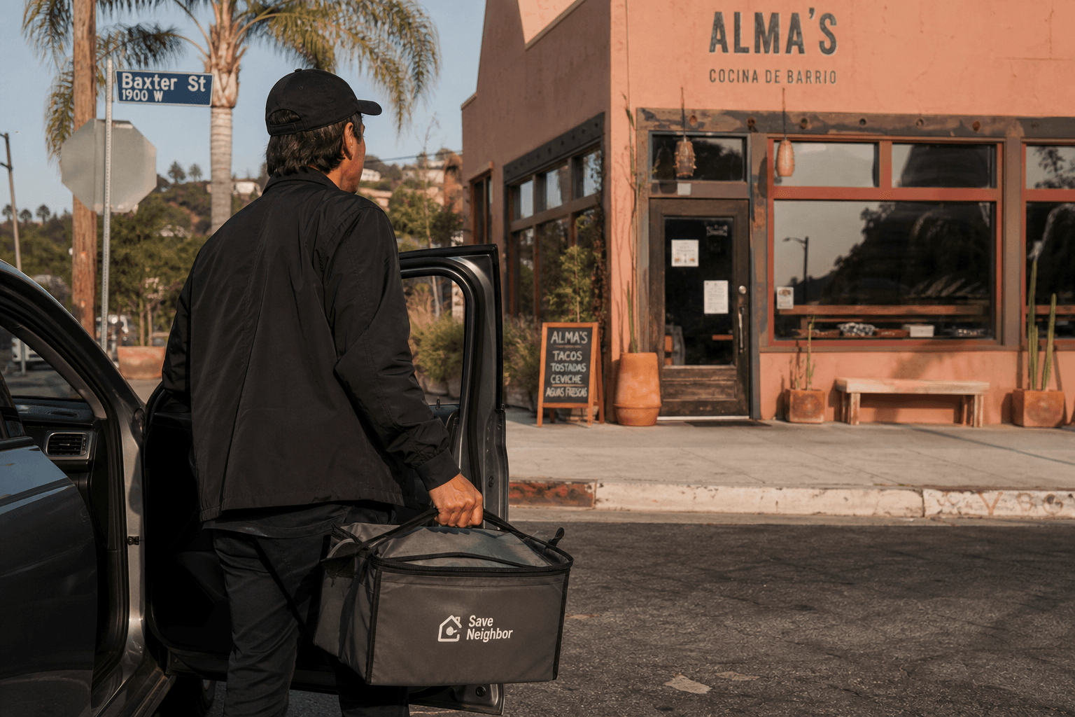 A delivery driver picks up an order from a neighborhood restaurant on a quiet Los Angeles street lined with palm trees.